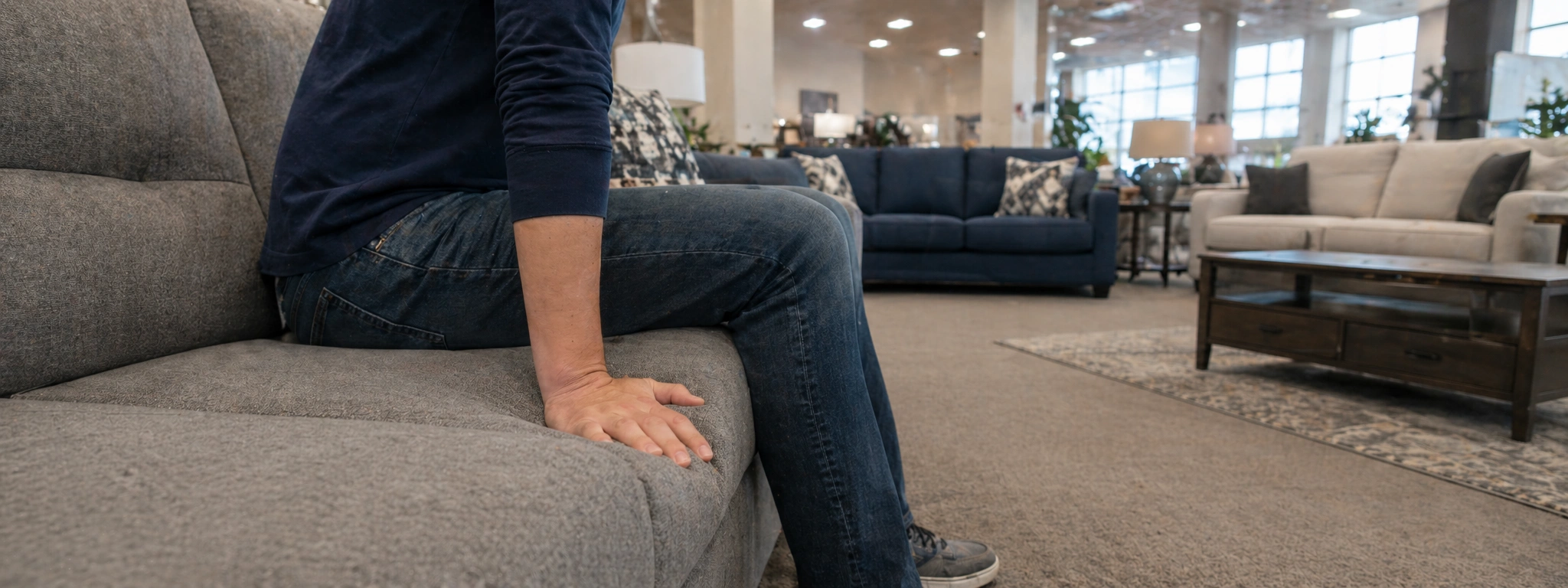 Person testing sofa cushion firmness by sitting and pressing with hands in a furniture showroom